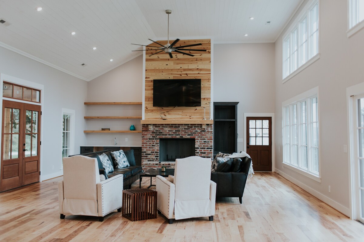 A spacious living room features large windows allowing ample natural light. A brick fireplace is flanked by wooden shelves. Two light-colored armchairs are positioned across from a dark coffee table and a flat-screen television mounted above the fireplace. Wood flooring creates a warm atmosphere.