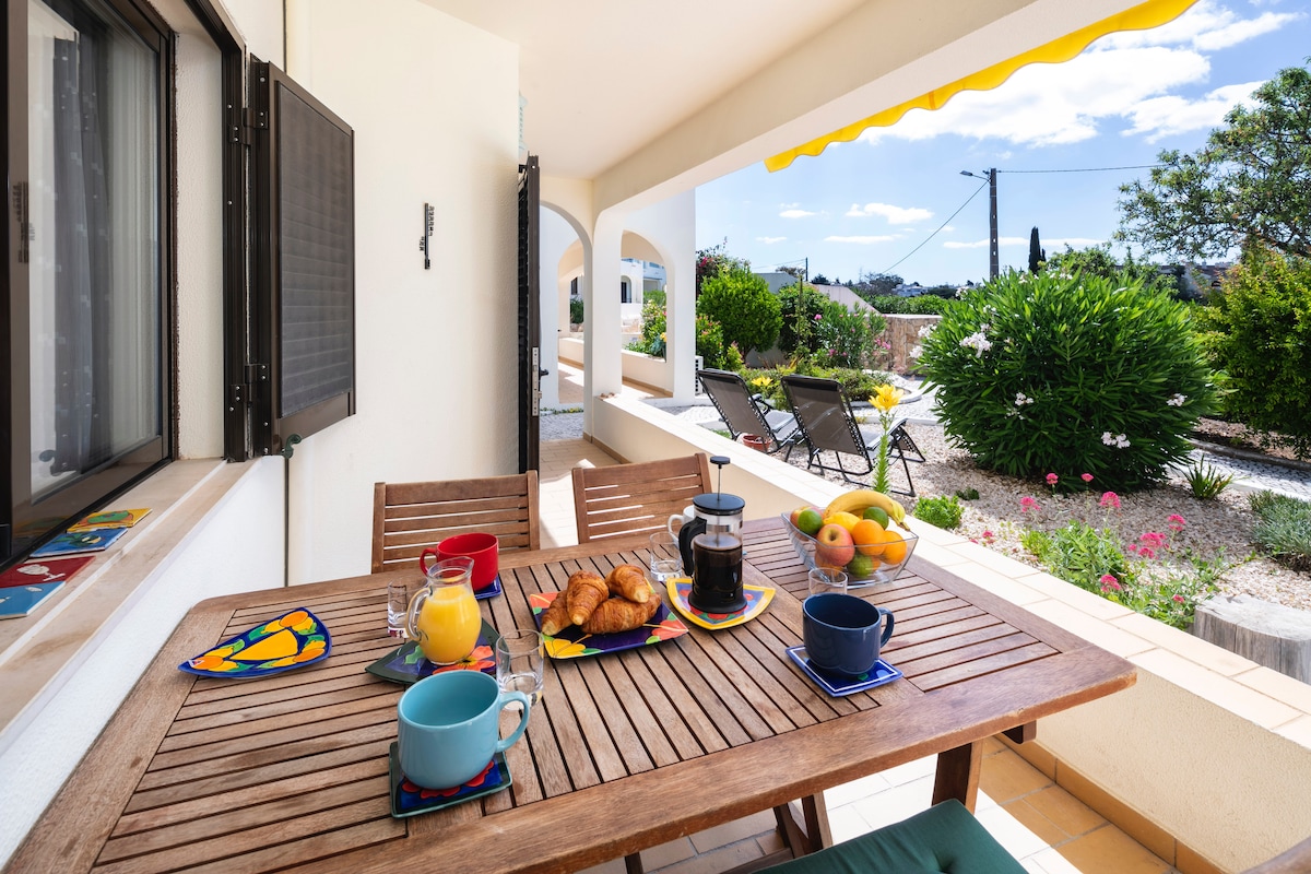 An outdoor dining area features a wooden table set with colorful cups, a coffee pot, fresh fruits, and pastries. A shaded terrace is complemented by reclining chairs and a vibrant awning, with well-maintained greenery visible in the background.