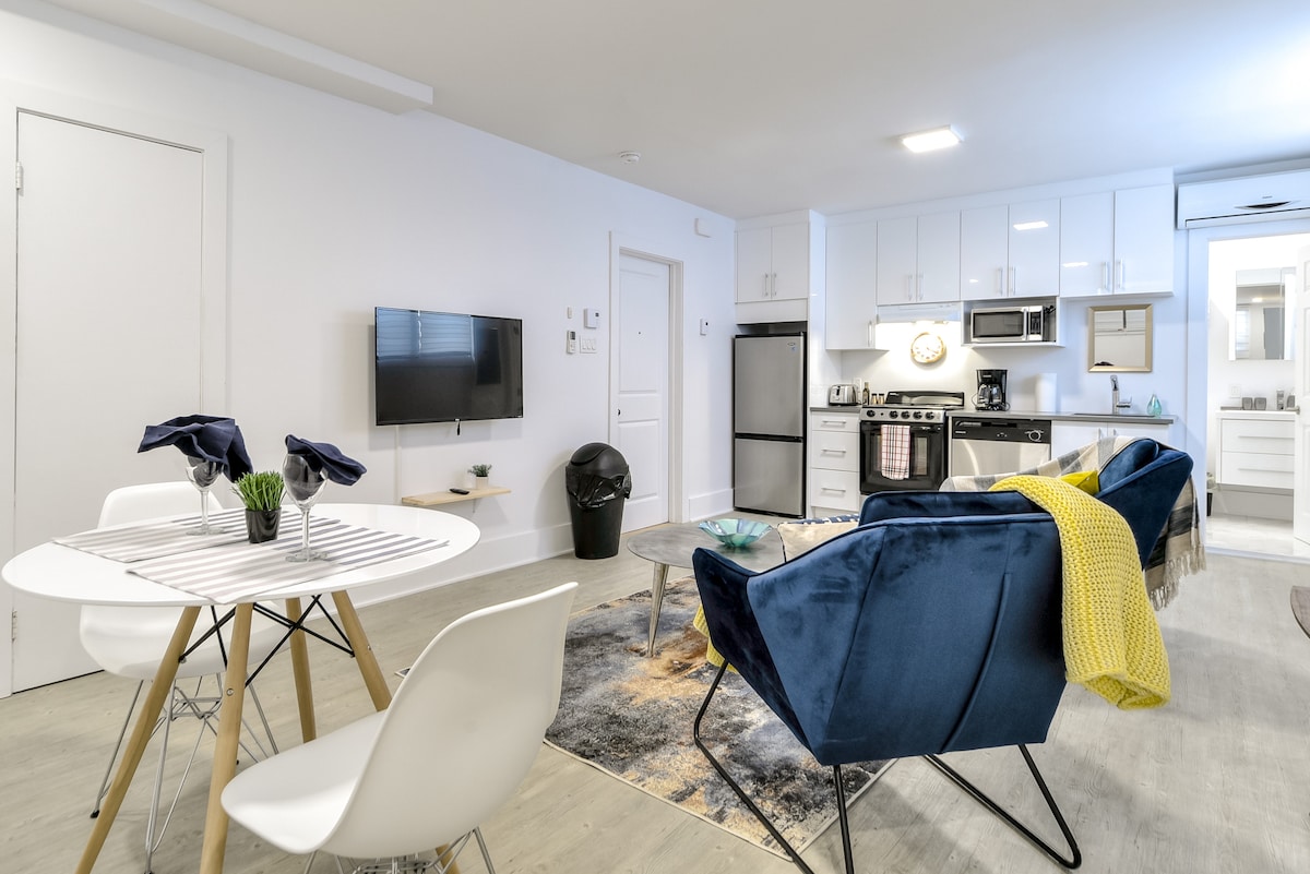 A bright living area combines a kitchen, dining space, and lounge, featuring modern appliances and furnishings. A round dining table is set with dark blue napkins, while a cozy blue chair with a yellow throw is positioned nearby. A large window allows ample natural light in.