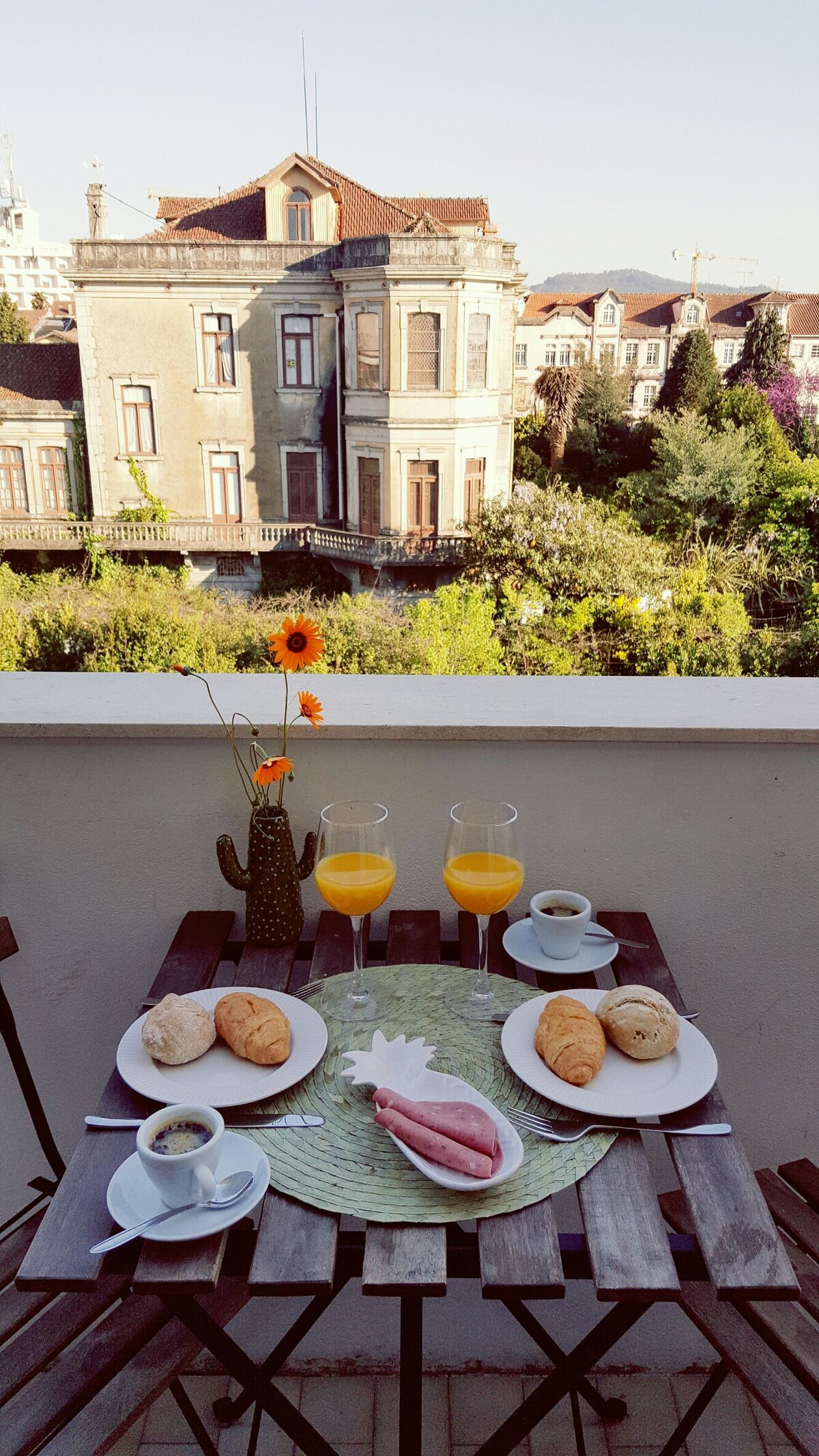 A small outdoor balcony features a round table set for breakfast, with two glasses of orange juice, two cups of coffee, and various pastries. A cactus and a flower in a vase adorn the table. The view includes a historic building and lush greenery.