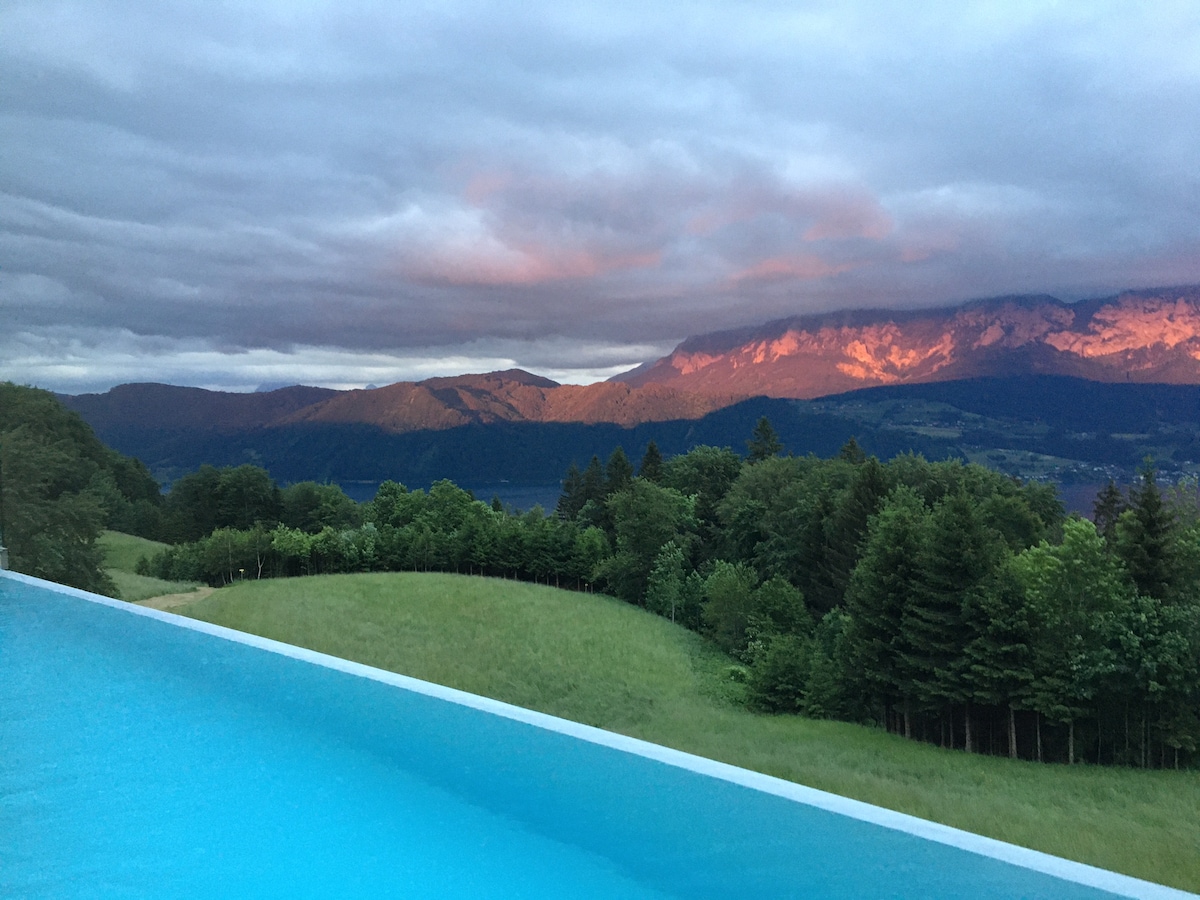 An expansive view showcases a tranquil outdoor pool with soft blue water, reflecting the dramatic sky. Nearby, lush green hills and dense forests are visible, while distant mountains are illuminated by the warm light of sunset, creating a serene natural backdrop.