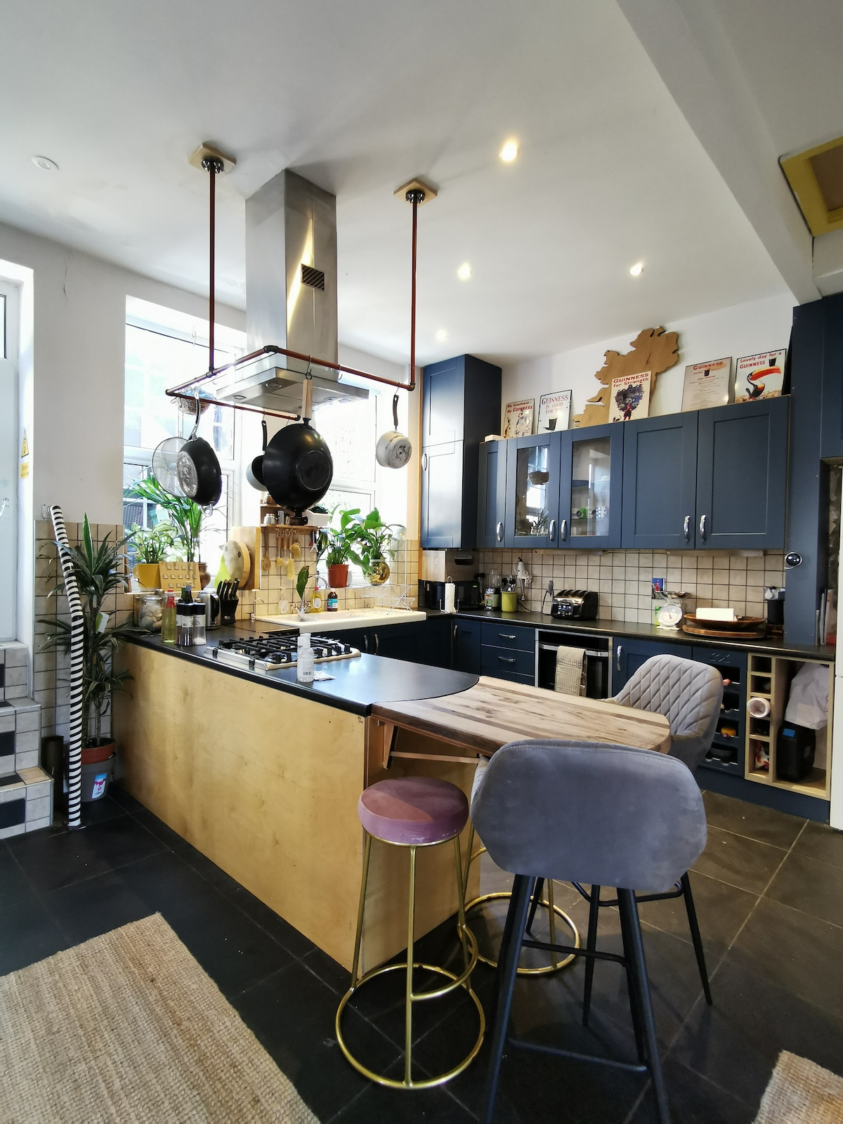 The spacious kitchen features a large central island with stylish stools, complemented by dark cabinetry and light tiling. Cookware hangs above the island, while plants add a touch of greenery. Natural light streams through large windows, enhancing the open feel of the room.