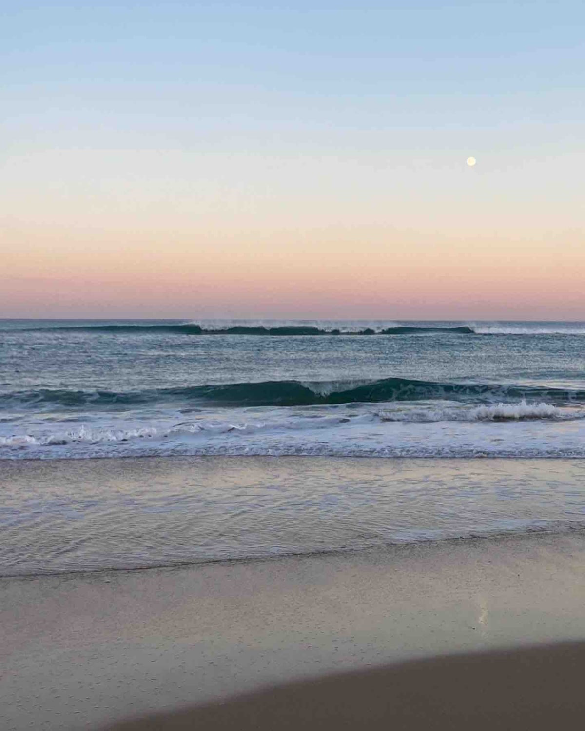 The image captures a serene beach scene at twilight, featuring gentle waves lapping against the shore. The sky transitions from soft pink to light blue, with the moon visible above the horizon, complementing the tranquil atmosphere.