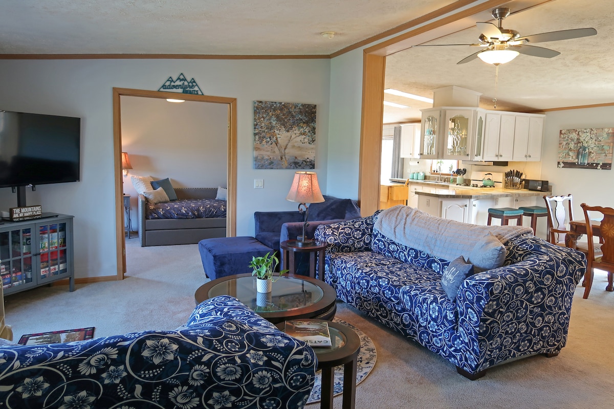 The living area features two large sofas in a floral pattern, complemented by a cozy armchair and small table. A flat-screen TV is mounted on the wall. A dining area is visible in the background, leading into a well-equipped kitchen.