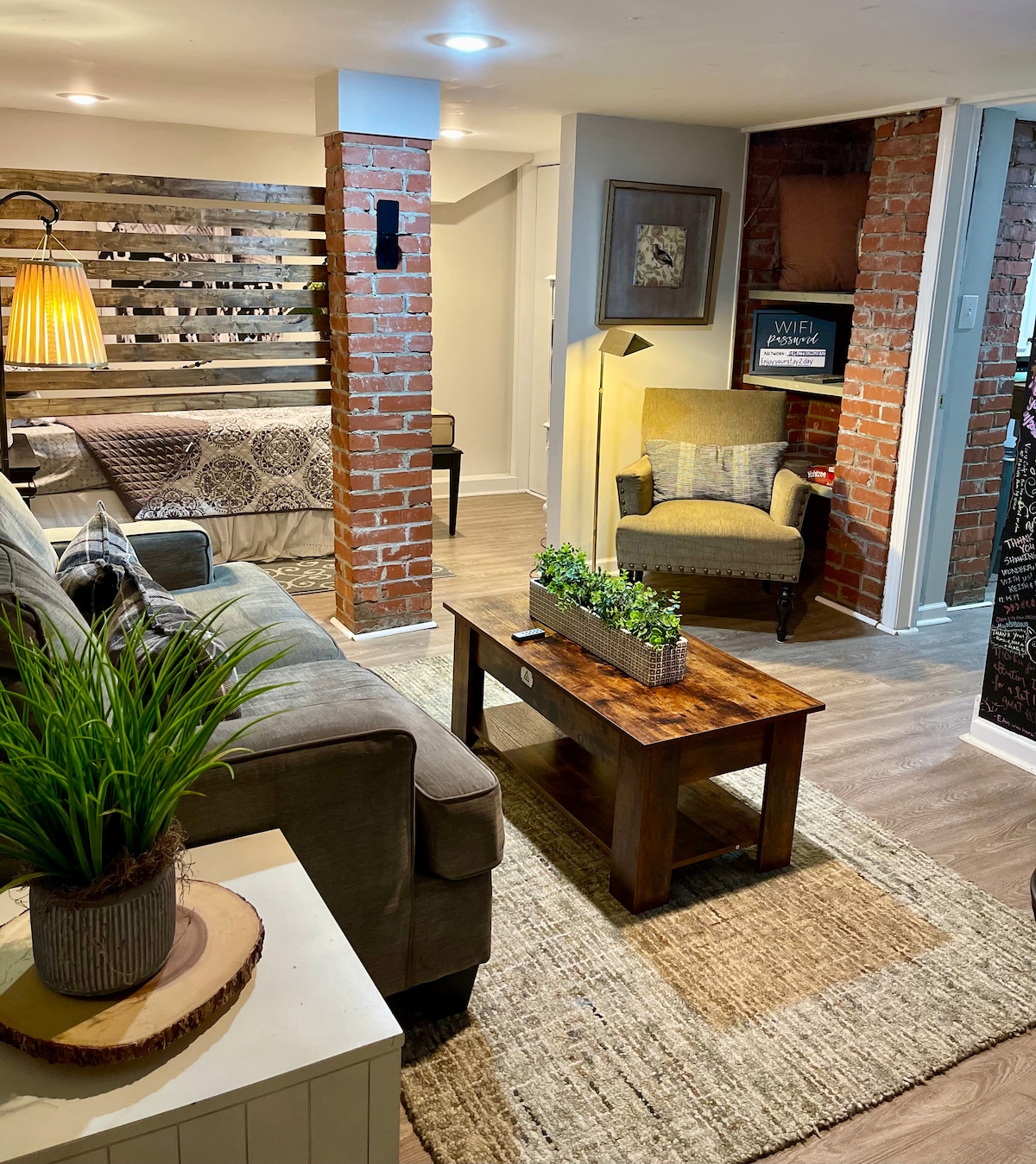 A living area features a comfortable grey sofa positioned beside a decorative reclaimed wood wall. A wooden coffee table sits atop a woven rug, accompanied by a small, stylish plant. Warm lighting illuminates the space, which is complemented by a corner accent chair.