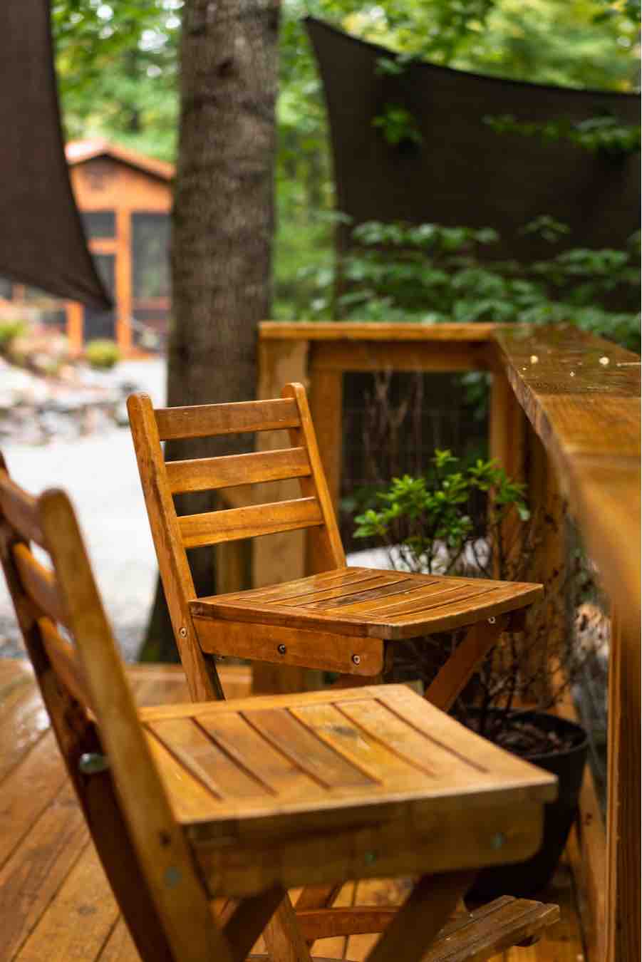Two wooden chairs are positioned on a deck surrounded by trees, creating a natural setting. The chairs are designed for comfort with slatted backs and seats. A wooden railing and greenery enhance the serene outdoor space.