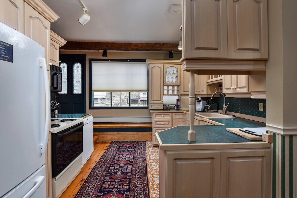 A spacious kitchen is equipped with white appliances, including a refrigerator and oven. Light wood cabinetry is complemented by a green countertop. A large window allows natural light, and a decorative area rug adds warmth to the wooden floor.