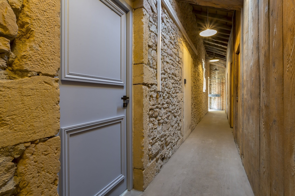A narrow corridor features a blend of stone and wooden walls, creating a rustic charm. Soft lighting illuminates the space, while a solid door is visible at the left. The far end opens to an airy area with natural light, enhancing the inviting atmosphere.