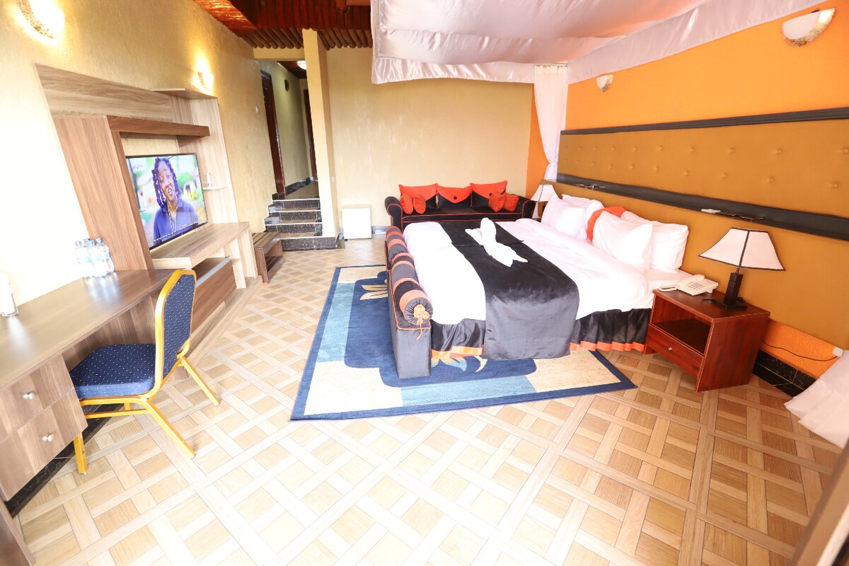 A spacious bedroom is presented, featuring a large bed with contrasting black and white bedding. A modern television is mounted on the wall opposite a wooden desk with a blue upholstered chair. The floor is adorned with a patterned tile, adding a touch of warmth to the space.