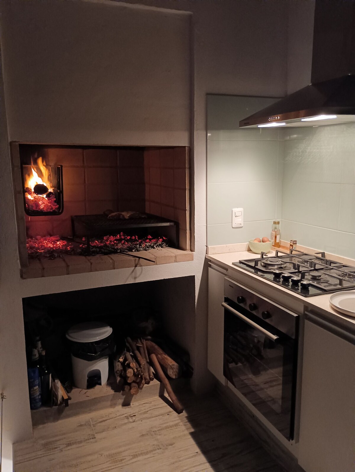 The kitchen area features a built-in grill with an open flame and glowing embers, providing a traditional cooking option. A modern gas stove with four burners complements the setup. Additional storage and firewood are neatly organized beneath the grill area.