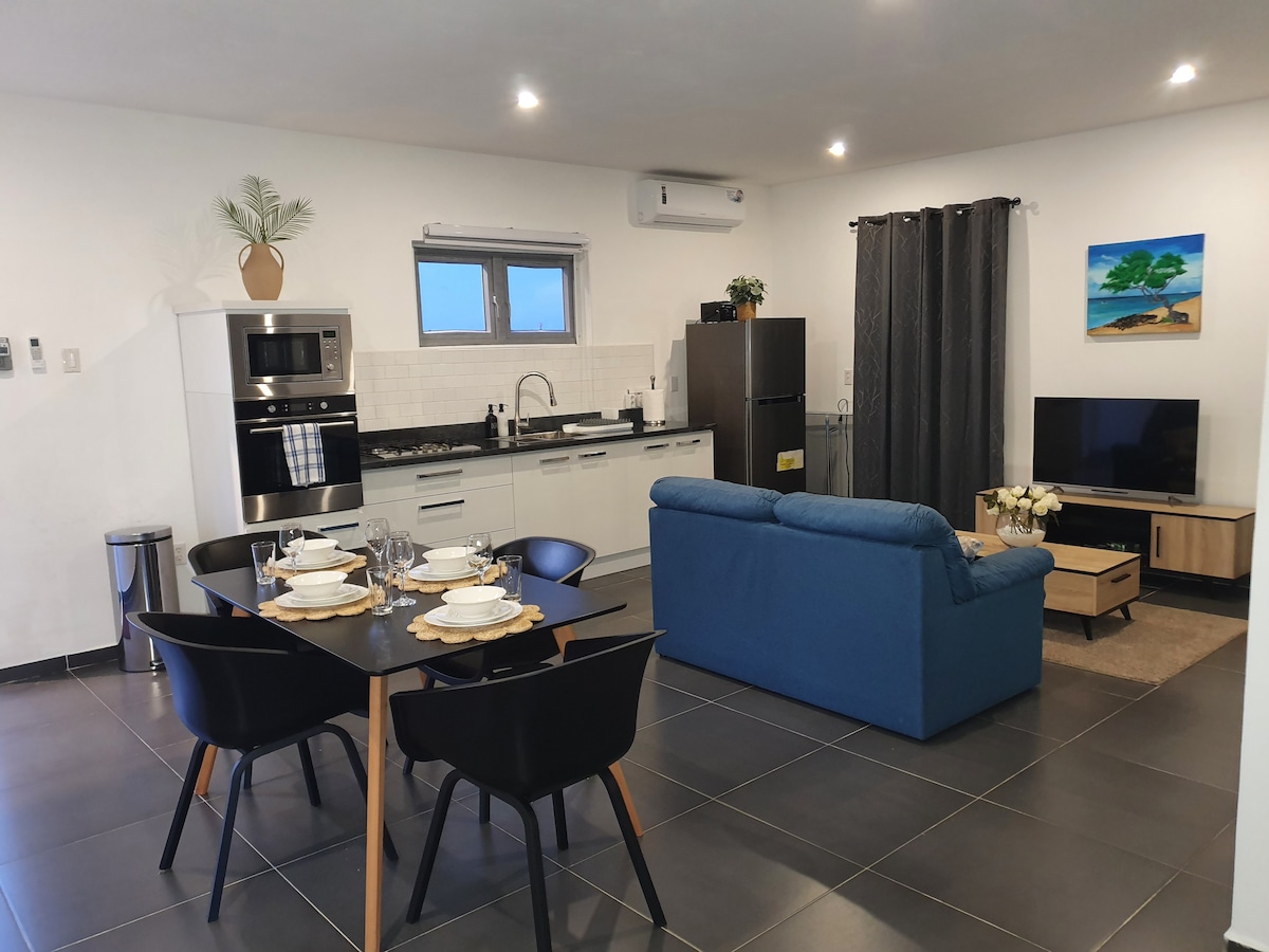 An open-plan living area is presented, featuring a kitchenette with modern appliances, a dining table set for four, and a comfortable blue sofa. A flat-screen TV is positioned on a wooden stand, while artwork and plants enhance the space. Natural light enters through a window.