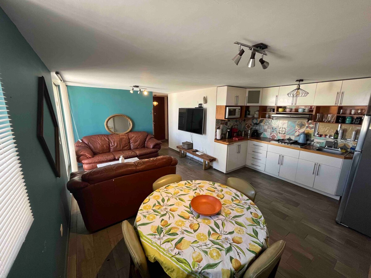 A well-lit living space is combined with a dining area, featuring a round table adorned with a lemon-patterned tablecloth. A brown leather couch is positioned opposite a modern kitchen, equipped with cabinets and appliances. Large windows provide natural light, enhancing the room's ambiance.