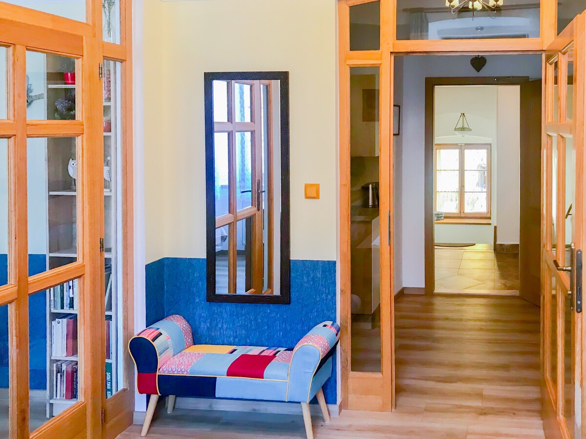 A welcoming entryway features wooden doors leading to an open space. A colorful bench with a patchwork design is positioned below a full-length mirror. Light-colored walls contrast with a blue accent wall, while warm wood flooring adds a pleasant touch.