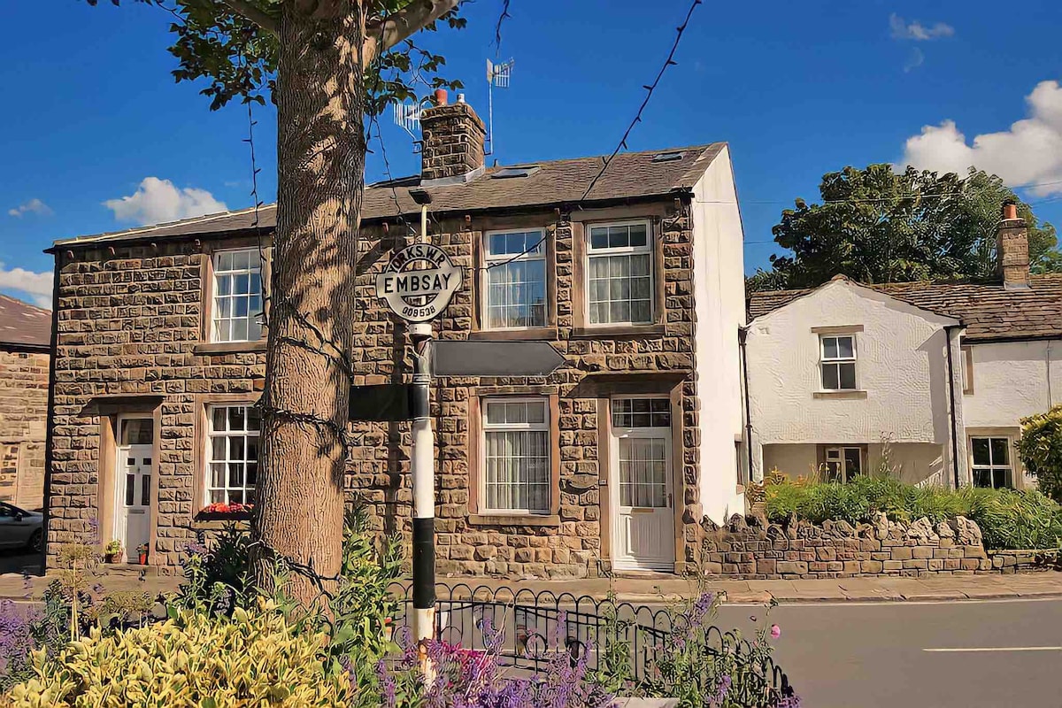 The exterior of Lovejoy cottage showcases stone walls and traditional architecture, with a well-maintained front garden highlighting colorful flowers. A street sign indicating 'Embsay' is positioned in front, surrounded by decorative fencing. The cottage is situated in a peaceful village setting with clear blue skies above.