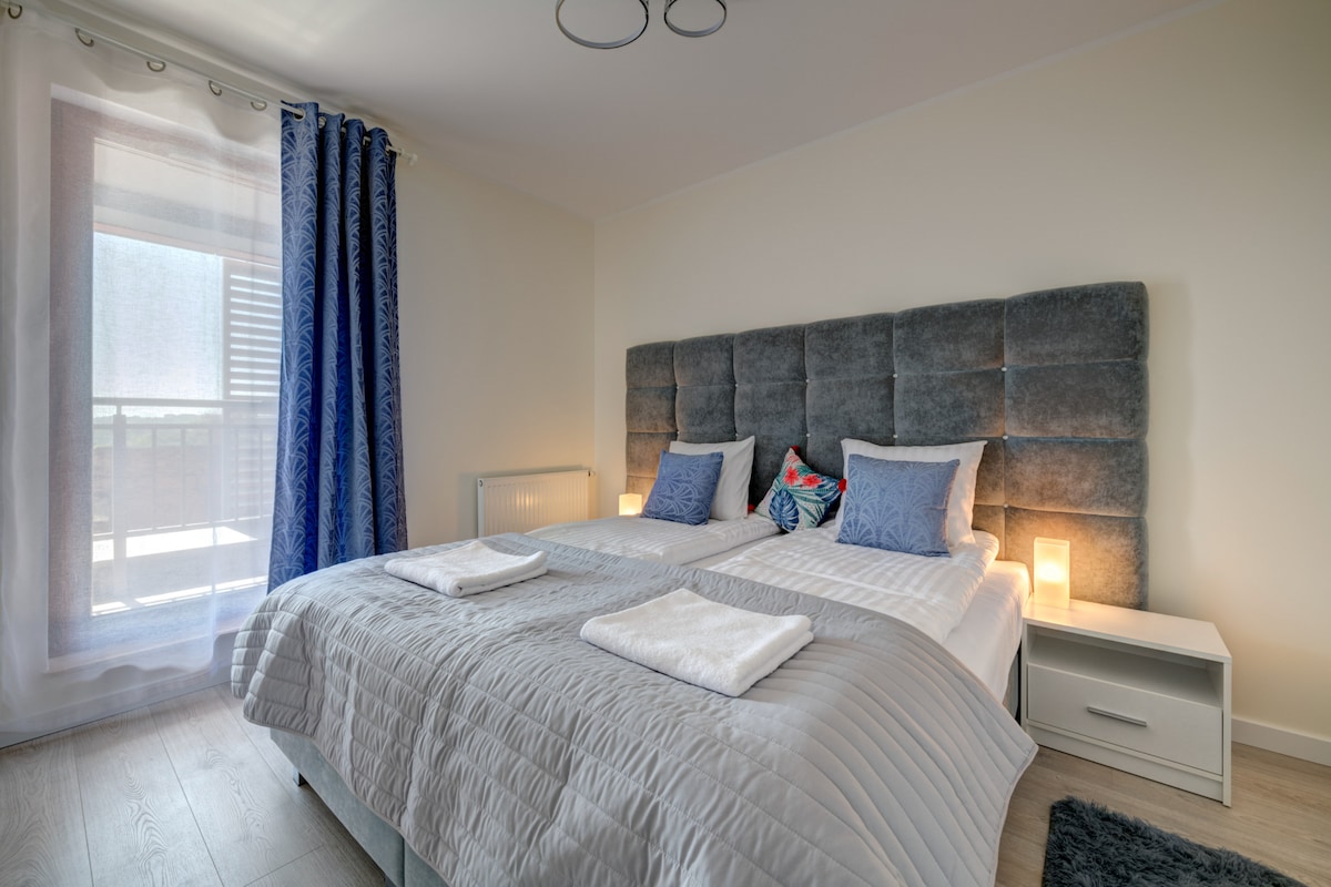 A spacious bedroom is furnished with two single beds, each adorned with soft blue pillows. Two bedside lamps provide gentle illumination, and a window draped with sheer curtains allows natural light to filter in. A modern grey upholstered headboard adds a contemporary touch.