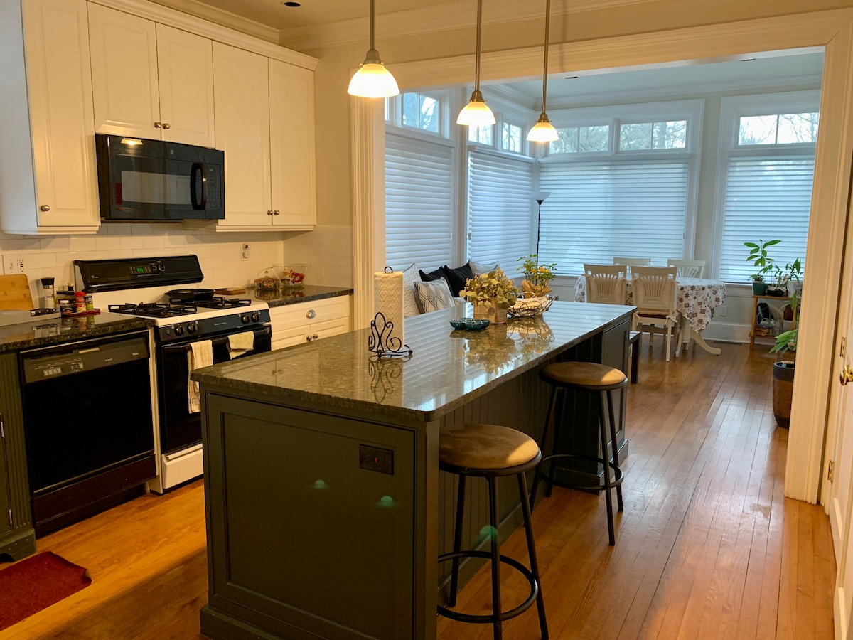 The kitchen features modern white cabinetry and a spacious island with bar stools. A black stove and microwave are visible, along with a light granite countertop. Adjacent to the kitchen, a sunlit dining area is seen through windows, showcasing plants and a dining table set for meals.