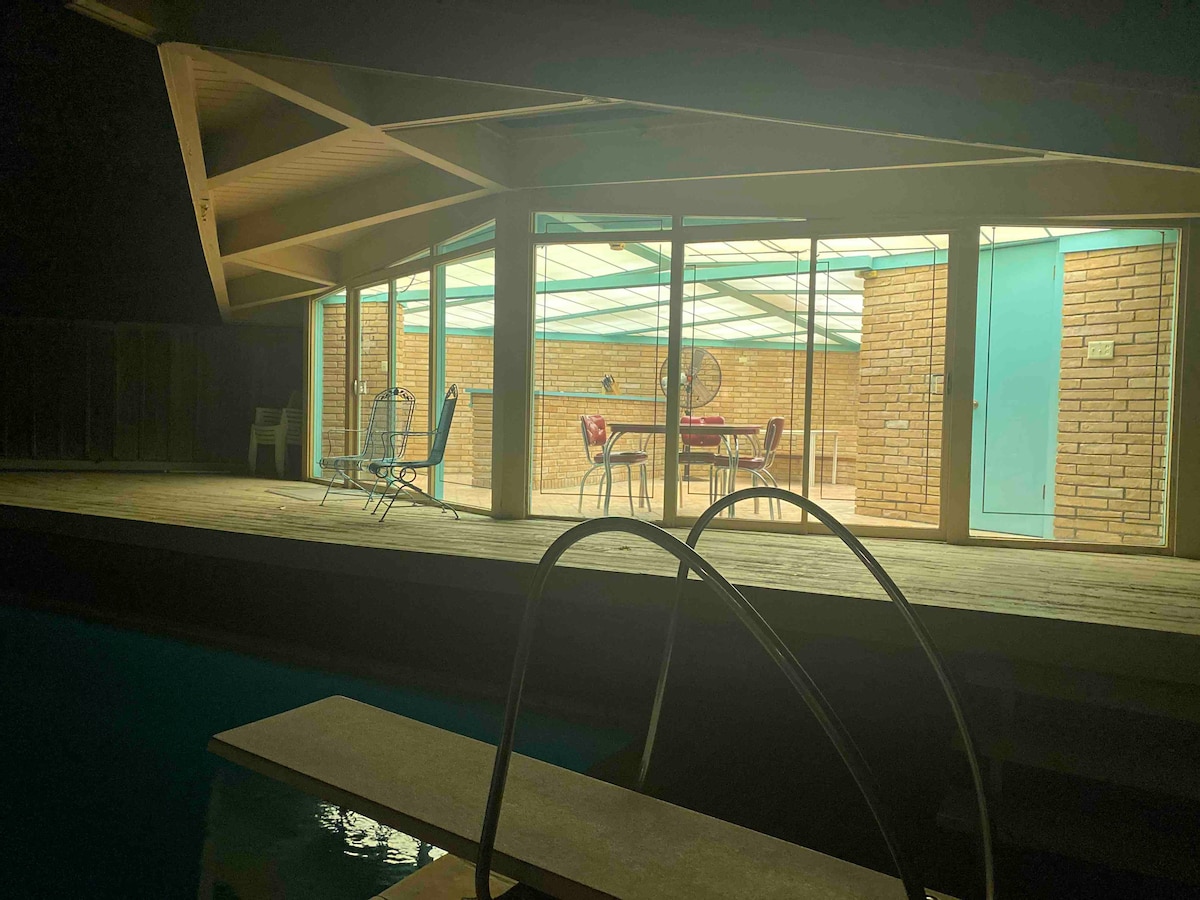 A poolside view captures a retro cabana illuminated at night, featuring large glass walls that reveal a dining area and seating. The inviting pool reflects soft light from the cabana, while a diving board extends into the water, enhancing the overall ambiance.