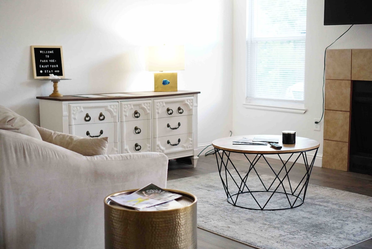 A spacious living area features a cozy sofa and a round coffee table with a geometric design. Natural light enters through the window, illuminating a decorative side table and an ornate cabinet, while a lighted sign offers a welcoming message.