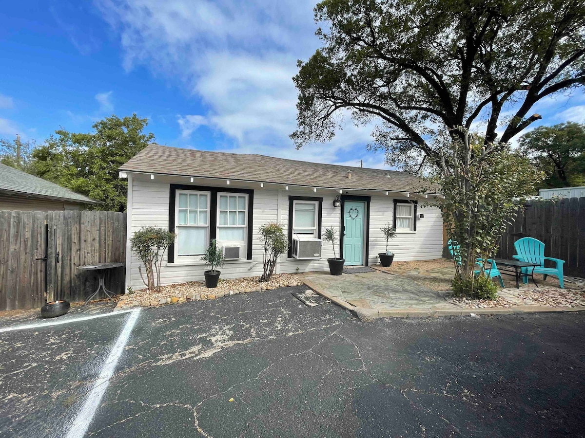 A guest house exterior is shown with a light-colored facade and a turquoise door. Two air conditioning units are placed on either side of the entrance. Green chairs are positioned in front, surrounded by small shrubs and a paved sitting area under a large tree.