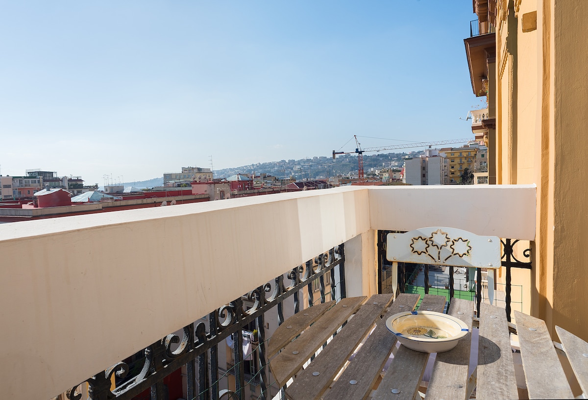 A balcony features wooden seating, offering a view of the sea and the coastline. A serene setting is provided by the open sky, with a plate of food placed on the table, inviting quiet moments outdoors.