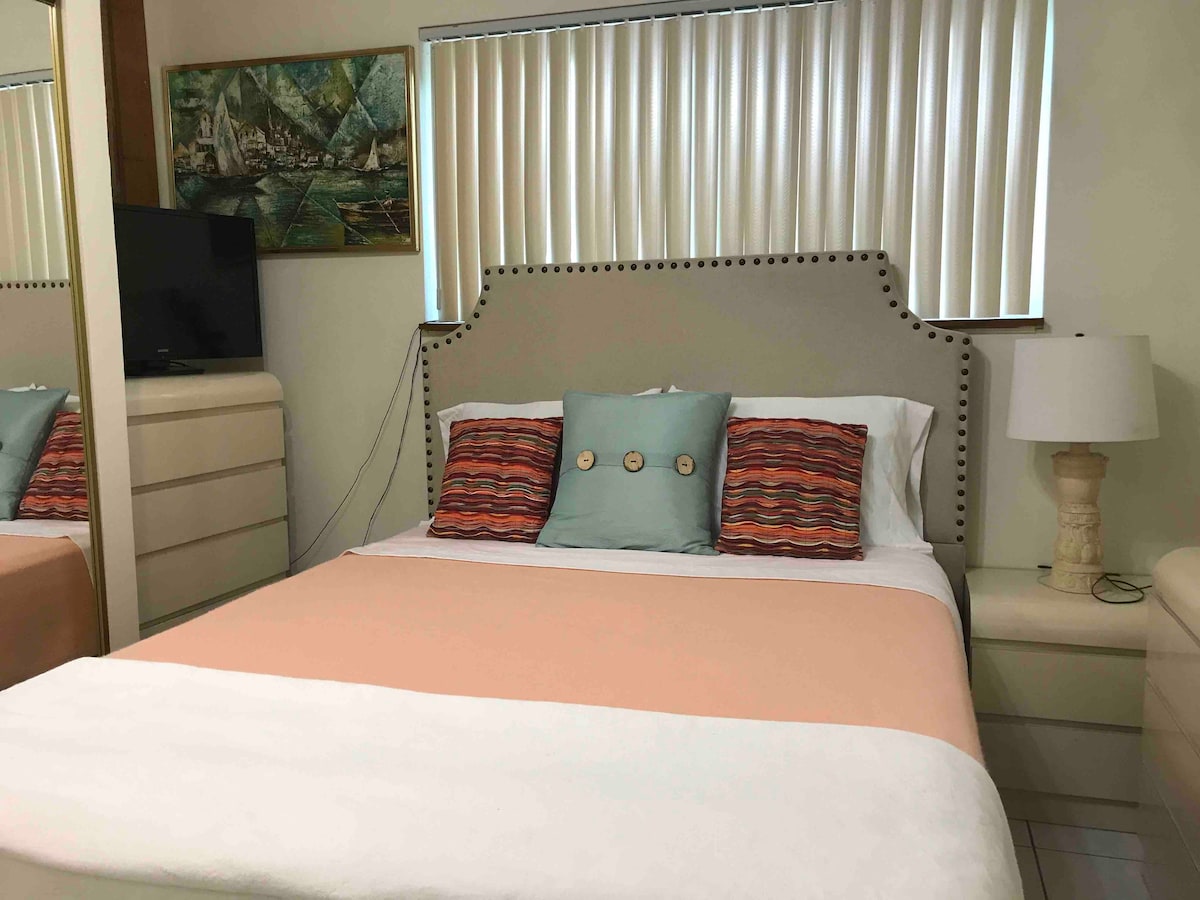 A comfortable sleeping area features a spacious bed dressed in soft linens, complemented by a decorative throw and patterned pillows. A bedside lamp stands next to the bed, while a piece of wall art and a mirrored cabinet enhance the room's serene ambiance.