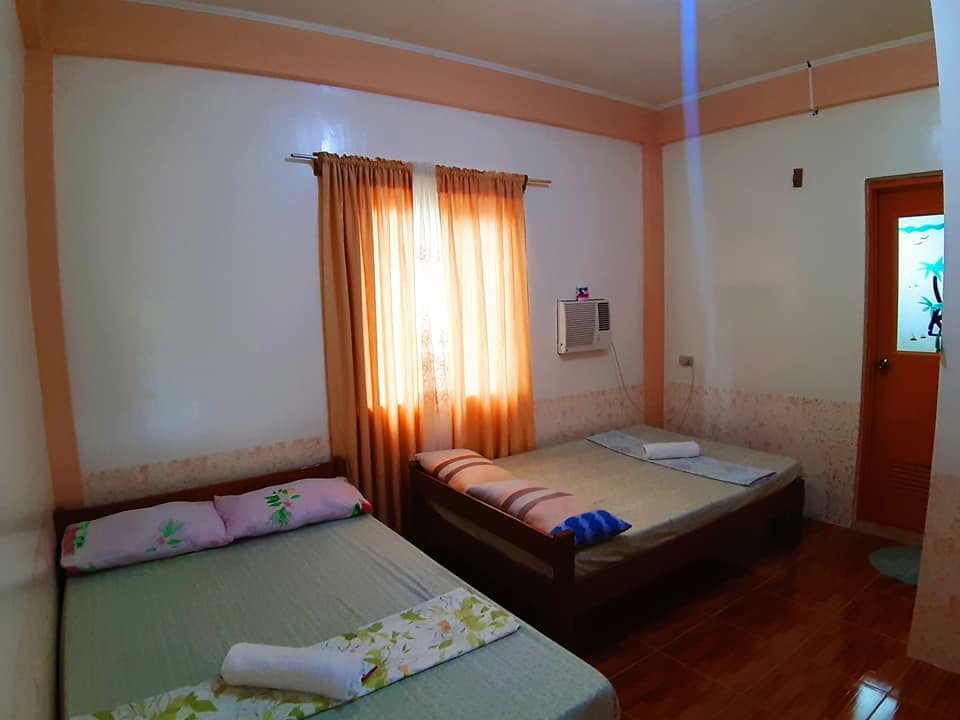 A well-kept bedroom accommodates two double beds positioned side by side. Soft curtains frame a window allowing natural light to fill the space. An air conditioning unit and a fan are present for comfort. Fresh linens and towels are neatly arranged on the beds.