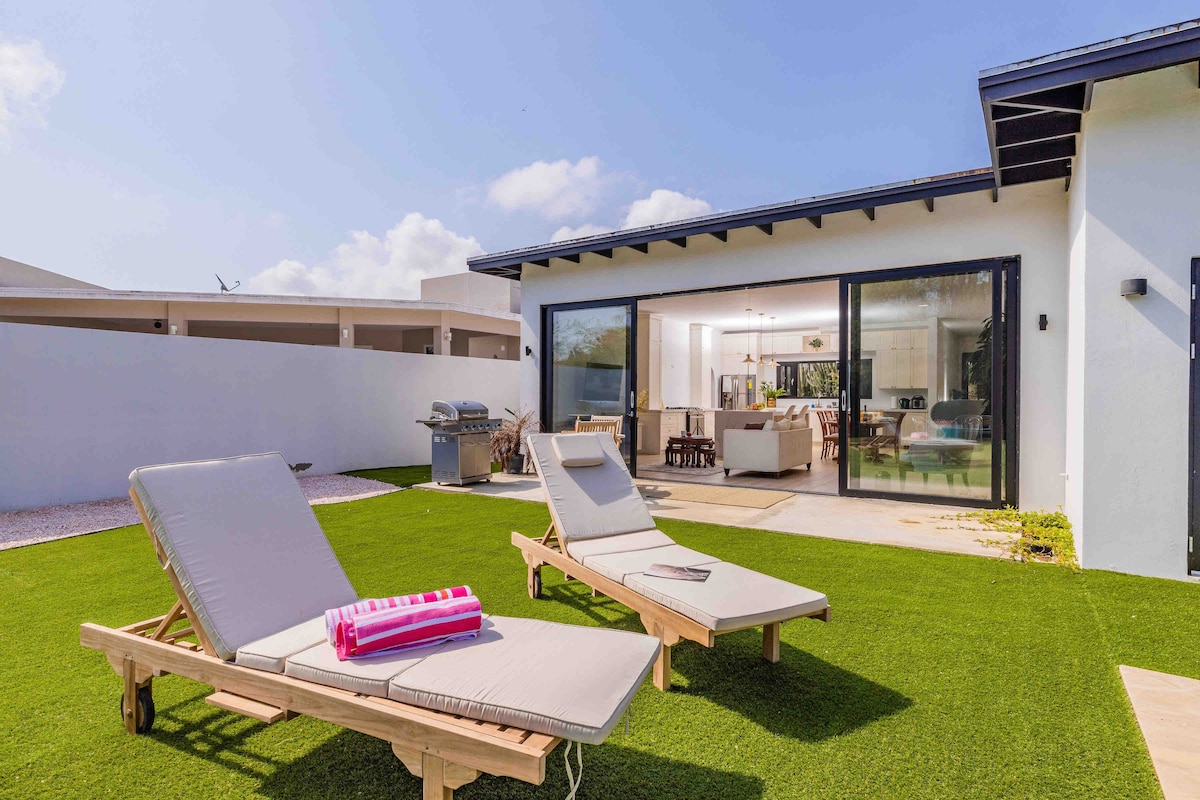 Two lounge chairs are positioned on a manicured grassy area, inviting relaxation. In the background, large sliding glass doors offer a glimpse into a spacious living area, while a barbecue grill is visible on the side. Bright blue skies complement the serene environment.
