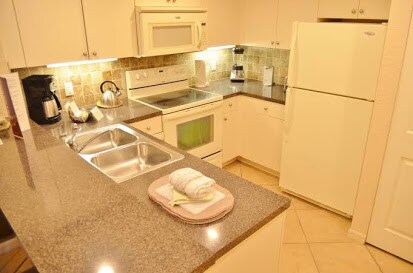 A functional kitchen is equipped with essential appliances, including a refrigerator, oven, and microwave. The counter features a double sink and is accented by a neatly arranged stack of towels. Warm lighting highlights the tile backsplash and neutral cabinetry, providing an efficient cooking space.