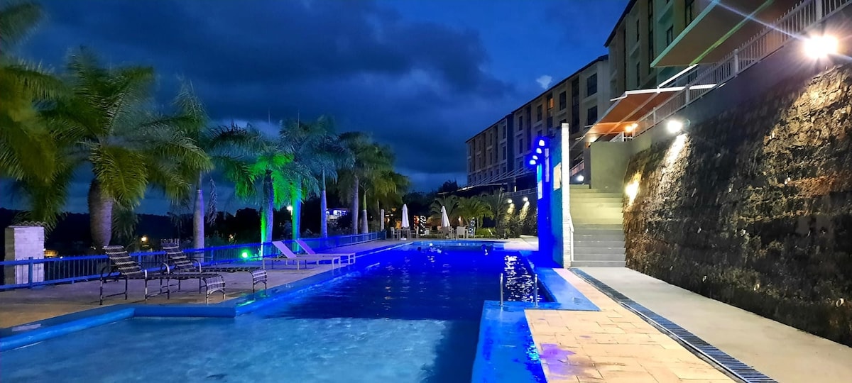 A pool area is illuminated by blue lights under a night sky, featuring a range of palm trees along the edge. Comfortable lounge chairs are positioned beside the water, with steps leading up to a multi-story building at the far end.