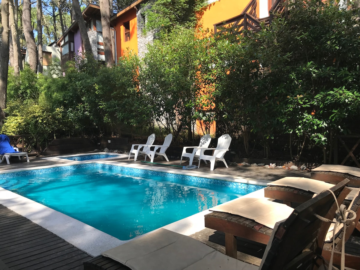A well-maintained outdoor pool area is surrounded by lush greenery, providing a serene setting. Several sun loungers are positioned beside the pool, inviting relaxation. The vibrant colors of the adjacent buildings contrast with the natural landscape, creating a welcoming ambiance.