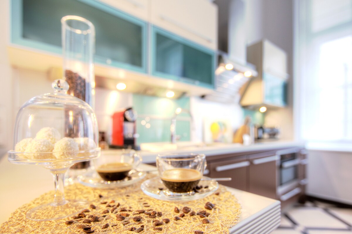 A modern kitchen is showcased with bright blue cabinetry and a spacious countertop. Two espresso cups are placed on a decorative mat surrounded by coffee beans. A glass cloche holds sweet treats, while under-cabinet lighting highlights the stylish design.