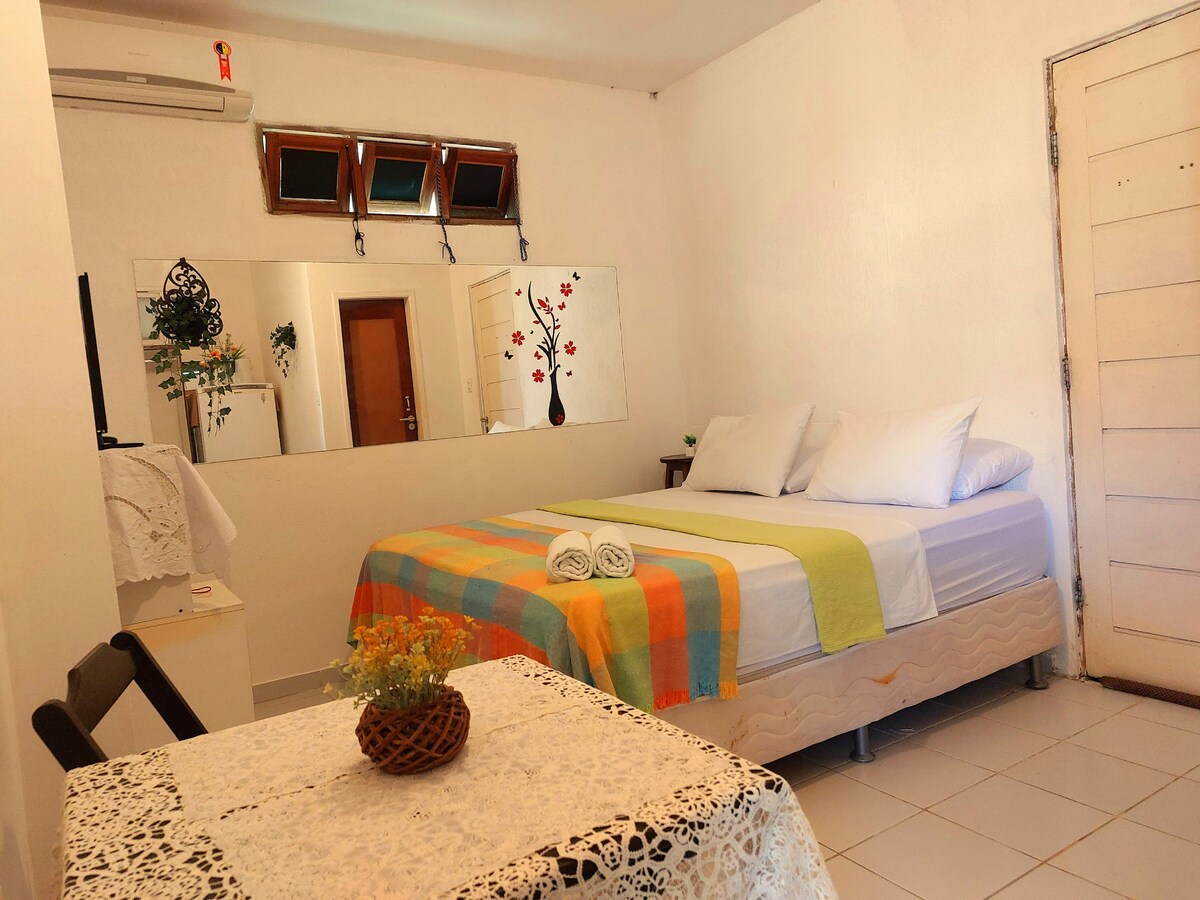 A spacious bedroom is featured, equipped with a large bed draped in light bedding. A mirror reflects the room's decor. A small table with a lace tablecloth is seen near the bed, while natural light is allowed through several windows at the top of the wall.