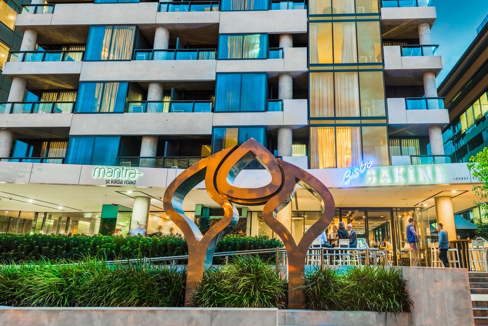 A contemporary apartment building features glass balconies and a blend of earthy tones in its facade. A modern sculpture stands prominently at the entrance, surrounded by lush green landscaping. Guests are seen enjoying the outdoor dining area under the soft glow of outdoor lighting.