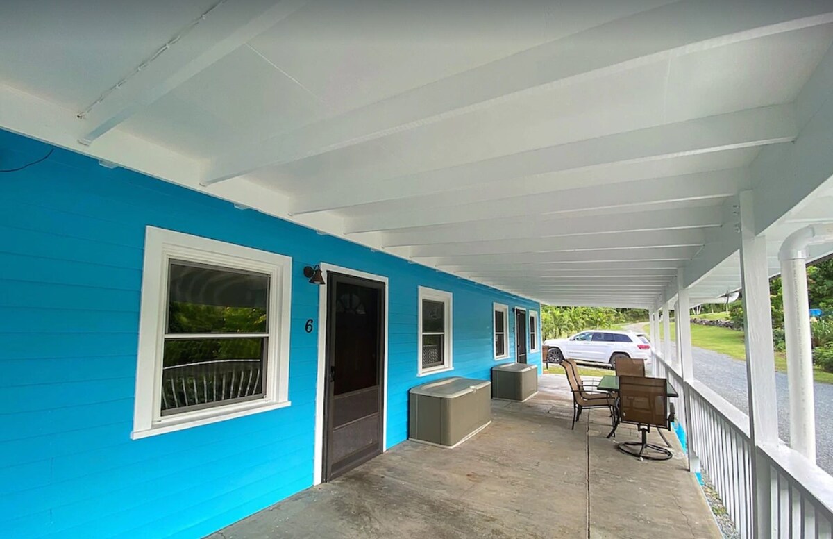 A covered porch is seen with bright blue walls and a concrete floor. Two chairs and a small table create a seating area, while large windows allow natural light into the space. Nearby, a vehicle is parked on the roadside.