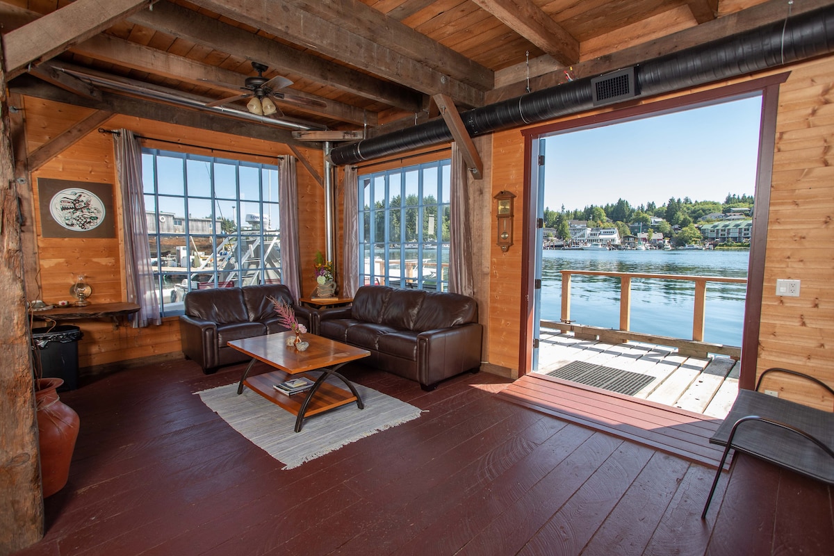 A cozy living area features large windows that provide views of the harbour, allowing natural light to fill the space. Timber beams offer a rustic charm, while a leather sofa and a wooden coffee table create a comfortable seating arrangement. A decorative rug adds warmth to the wooden floor.