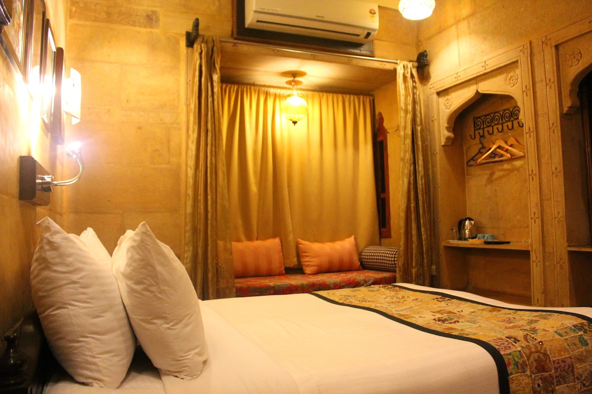 A cozy room features a spacious bed with plush pillows and a decorative bedspread. Natural light filters through the soft curtains, illuminating the traditional wall decor. A seating area with vibrant cushions and a closet with hangers is visible, complemented by modern amenities including an electric kettle.