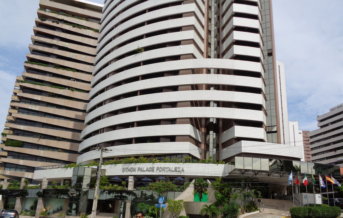 A modern hotel building is depicted, featuring a unique curved facade with horizontal stripes. The entrance is enhanced by greenery, with several flags displayed at the front. Multiple stories are visible, showcasing a blend of architecture and landscaping.