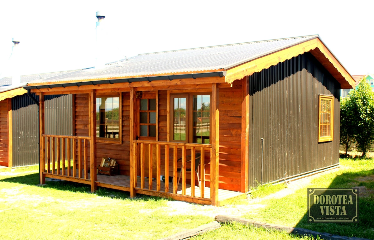 The exterior of a wooden cabin is showcased, featuring a combination of natural wood and dark paneling. A covered porch with a railing provides a welcoming entryway. Surrounding green grass complements the cabin's rustic charm.