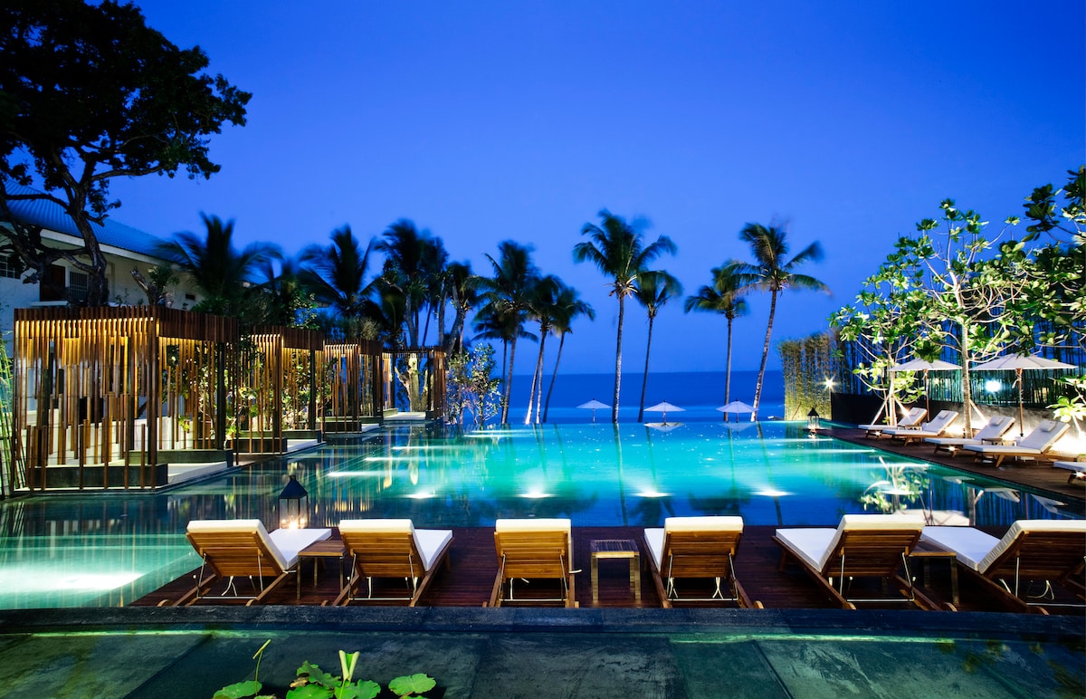An illuminated pool area is visible at dusk, surrounded by palm trees and lush greenery. Lounge chairs are arranged around the pool, reflecting in the water. The tranquil atmosphere is enhanced by the ocean in the background, adding a serene touch to the space.