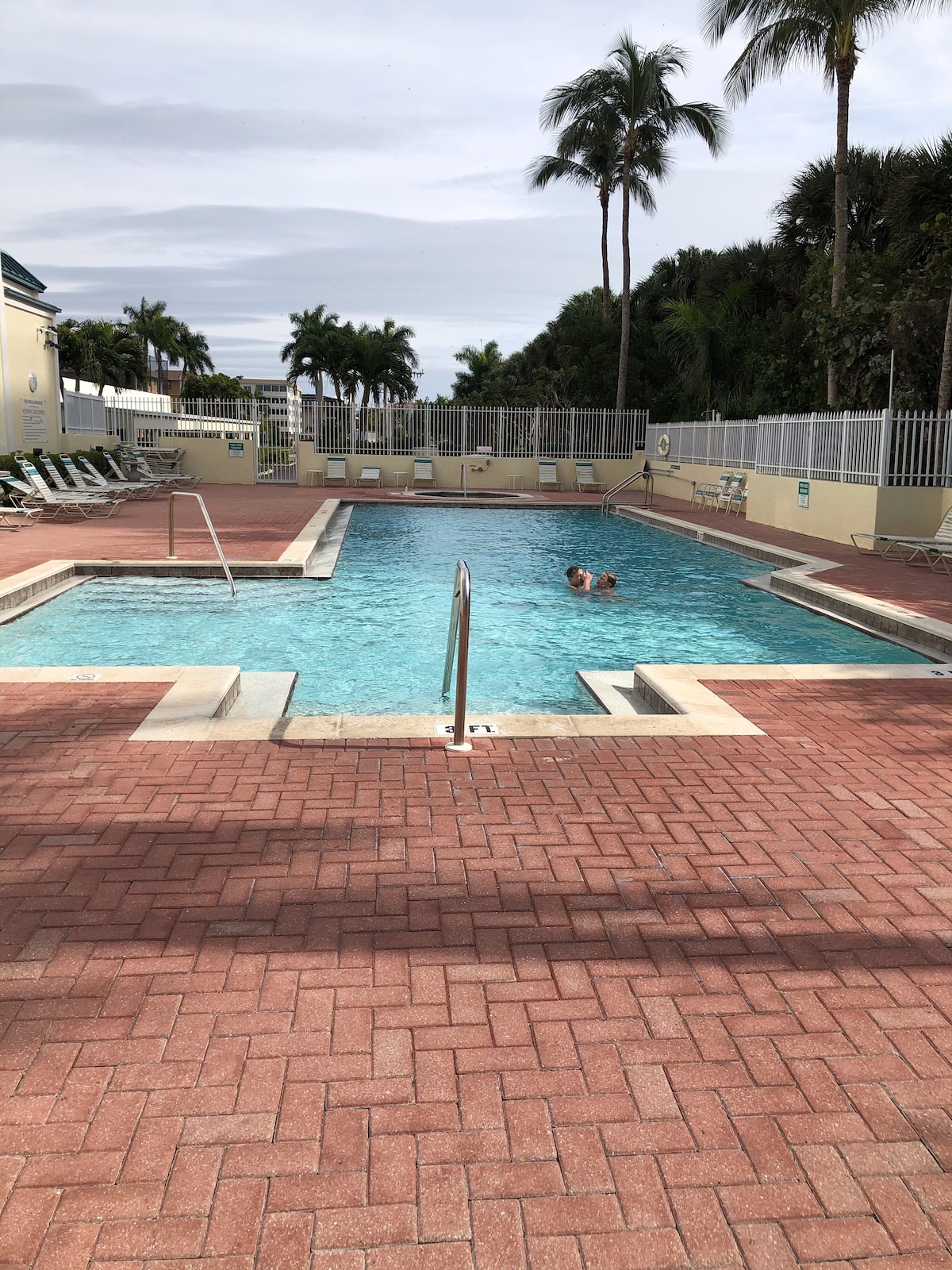 The image features a swimming pool framed by a brick patio, surrounded by lounge chairs under palm trees. A gentle slope leads to the shallow end, with stairs for entry. Two individuals can be seen enjoying the water in the inviting, clear pool.