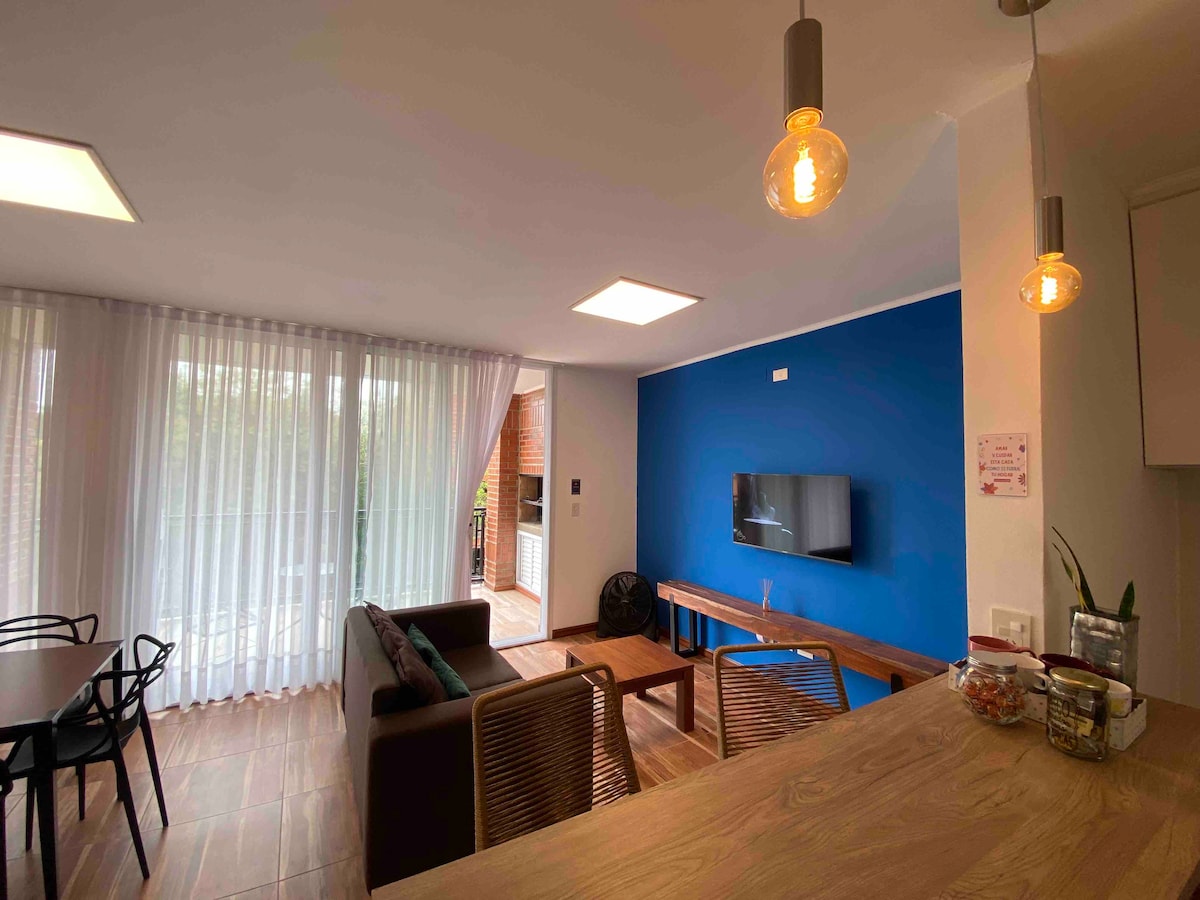 The living space is designed with a mix of contemporary and cozy elements. A dark sofa faces a wall-mounted television, complemented by natural light filtering through sheer curtains. A wooden table and chairs are positioned nearby, creating an inviting area for relaxation or dining.