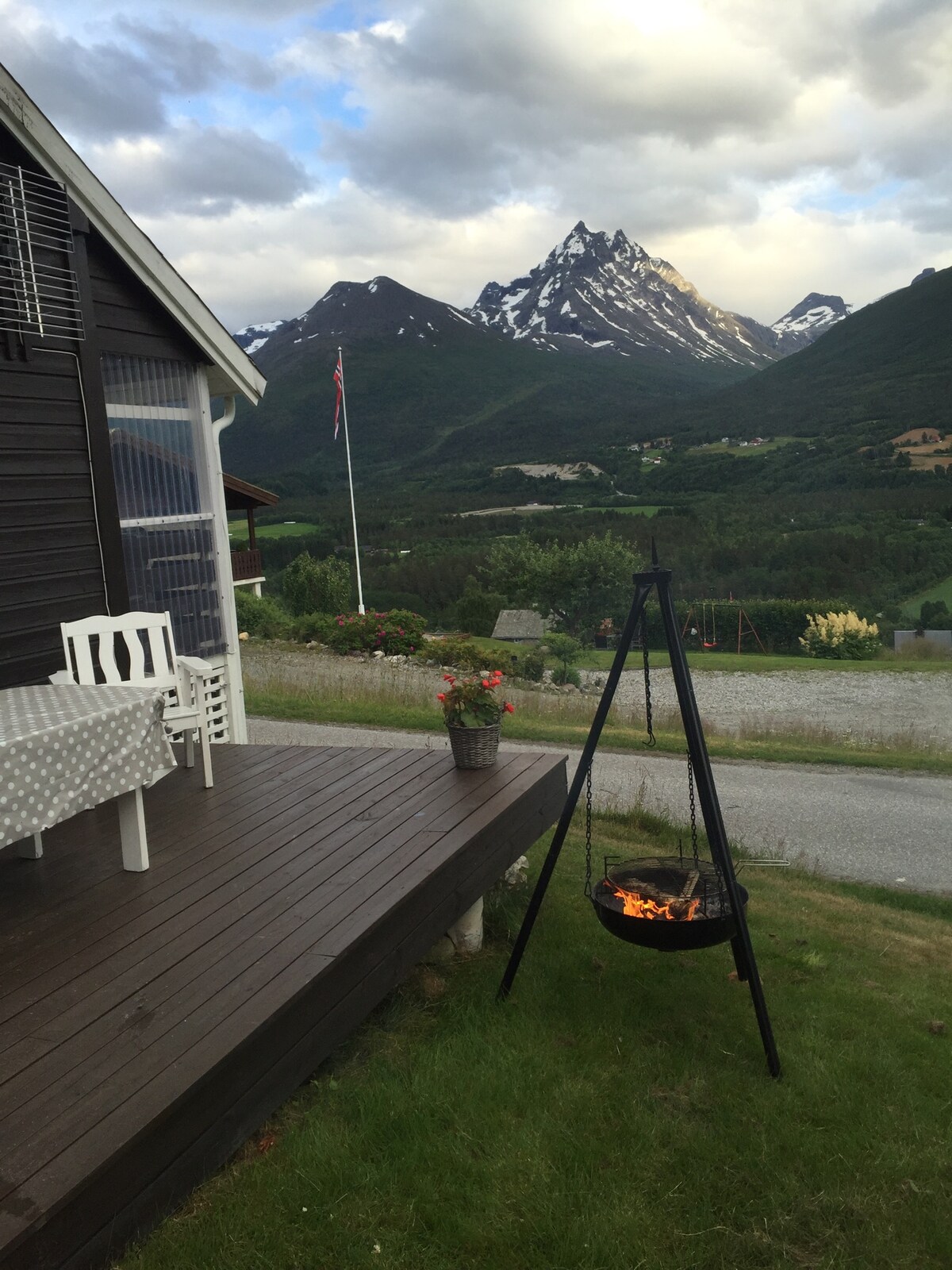 A wooden deck is showcased next to a dark exterior of the house, featuring a white dining set. A portable grill stands ready for use, with flames visible. In the background, snow-capped mountains rise against a cloudy sky, enhancing the natural scenery.