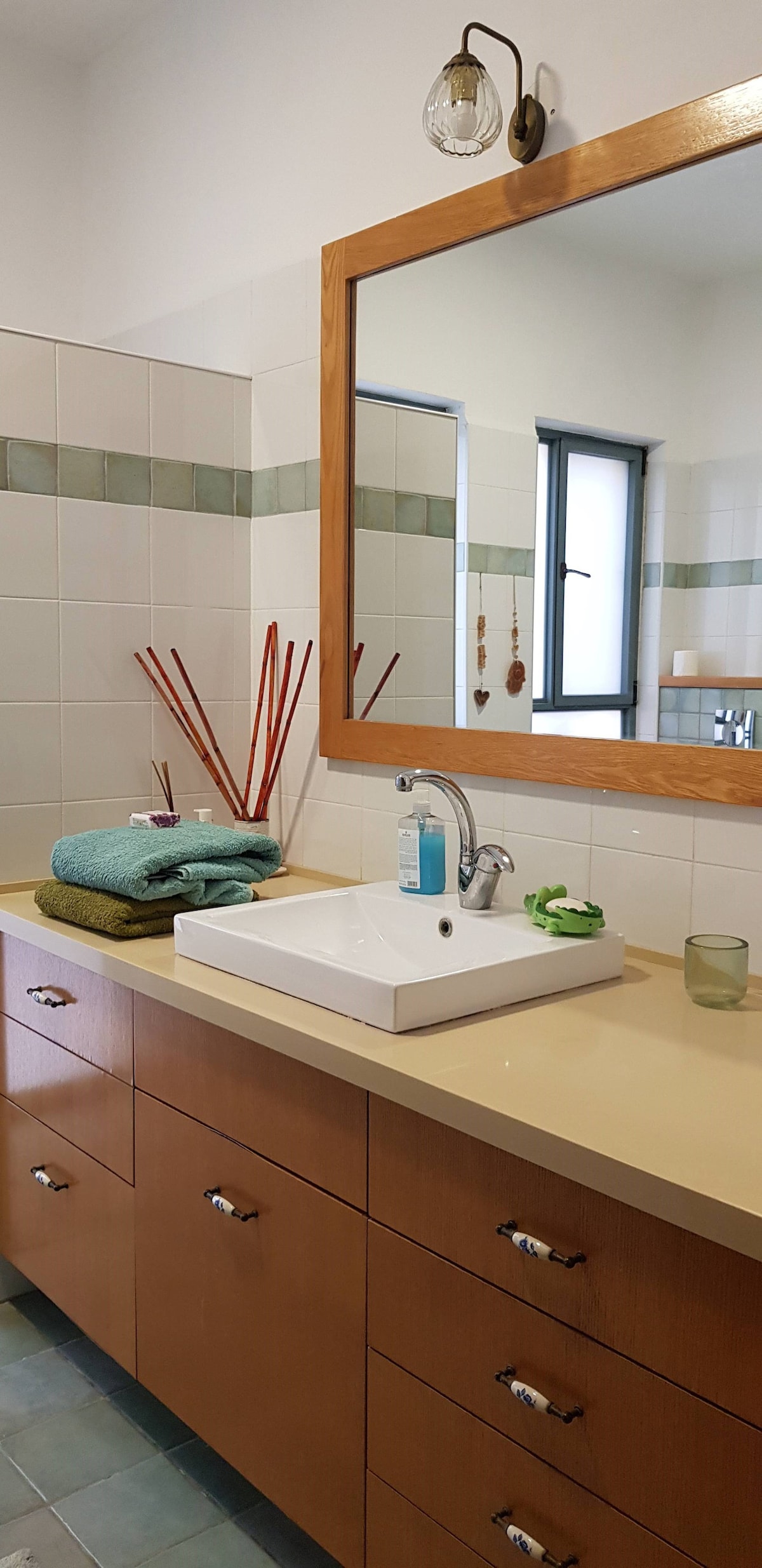 A well-lit bathroom features a large mirror above a modern sink with a minimalist design. Wooden cabinetry offers storage below, complemented by neatly arranged towels in a variety of colors. The space is enhanced by decorative elements like bamboo sticks and soft lighting.