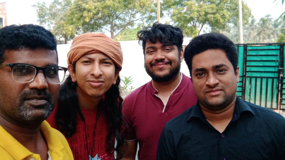 A group of four individuals stands outdoors, with trees and greenery visible in the background. The setting appears casual, and the people are dressed in various styles. Varying colors of clothing, including yellow, red, and black, contribute to the diverse visual elements.