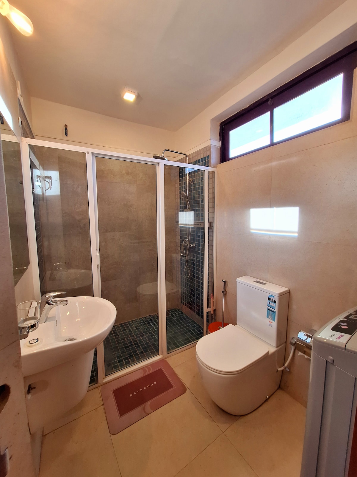 The bathroom features a glass shower cubicle, modern toilet, and a compact sink with a mirror. Natural light enters through a window, and a washing machine is placed nearby. Neutral tones and clean lines create a contemporary and functional space.