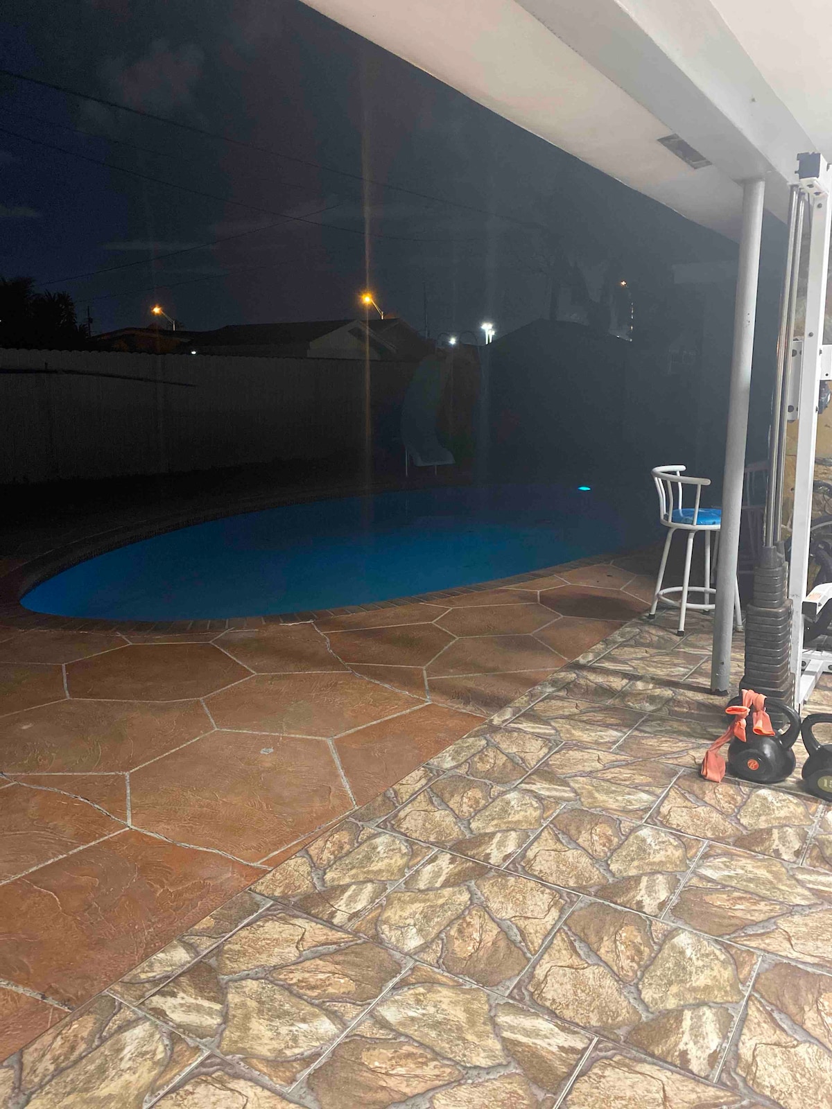A tranquil pool glows softly under the night sky, surrounded by carefully arranged stone tiles. A small bar stool is positioned nearby, while exercise equipment is placed against the wall, hinting at an active lifestyle. Soft outdoor lighting enhances the serene atmosphere.