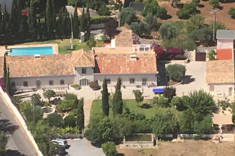 An aerial view of a spacious property features a traditional house with a tiled roof surrounded by greenery. A large swimming pool is visible to the left, while cozy outdoor seating areas and vibrant gardens add to the serene environment.