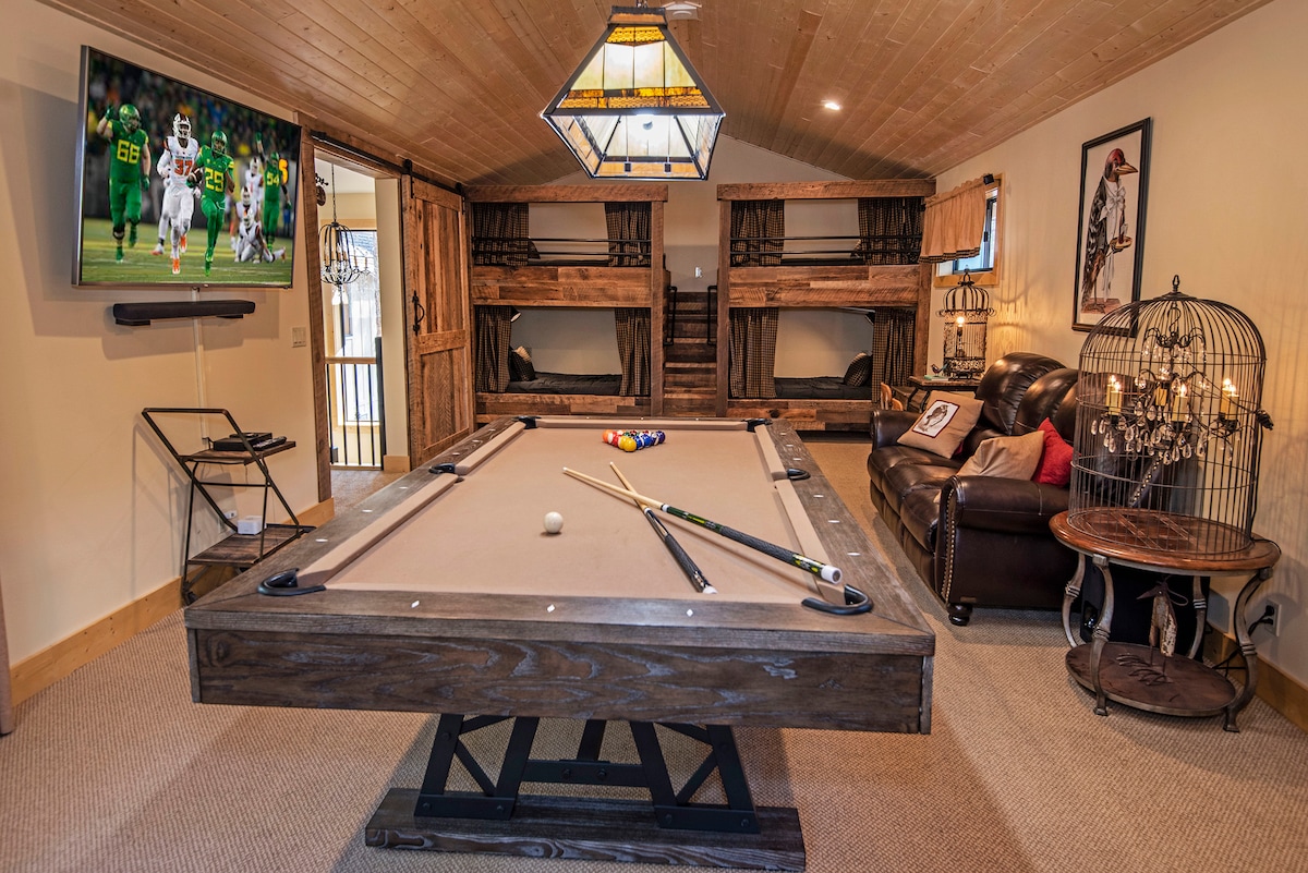 A recreational room features a spacious pool table positioned at the center under a wooden ceiling. Bunk beds are visible in the background, along with a comfortable seating area. A large television displays content mounted on the wall, complemented by soft lighting from an overhead fixture.