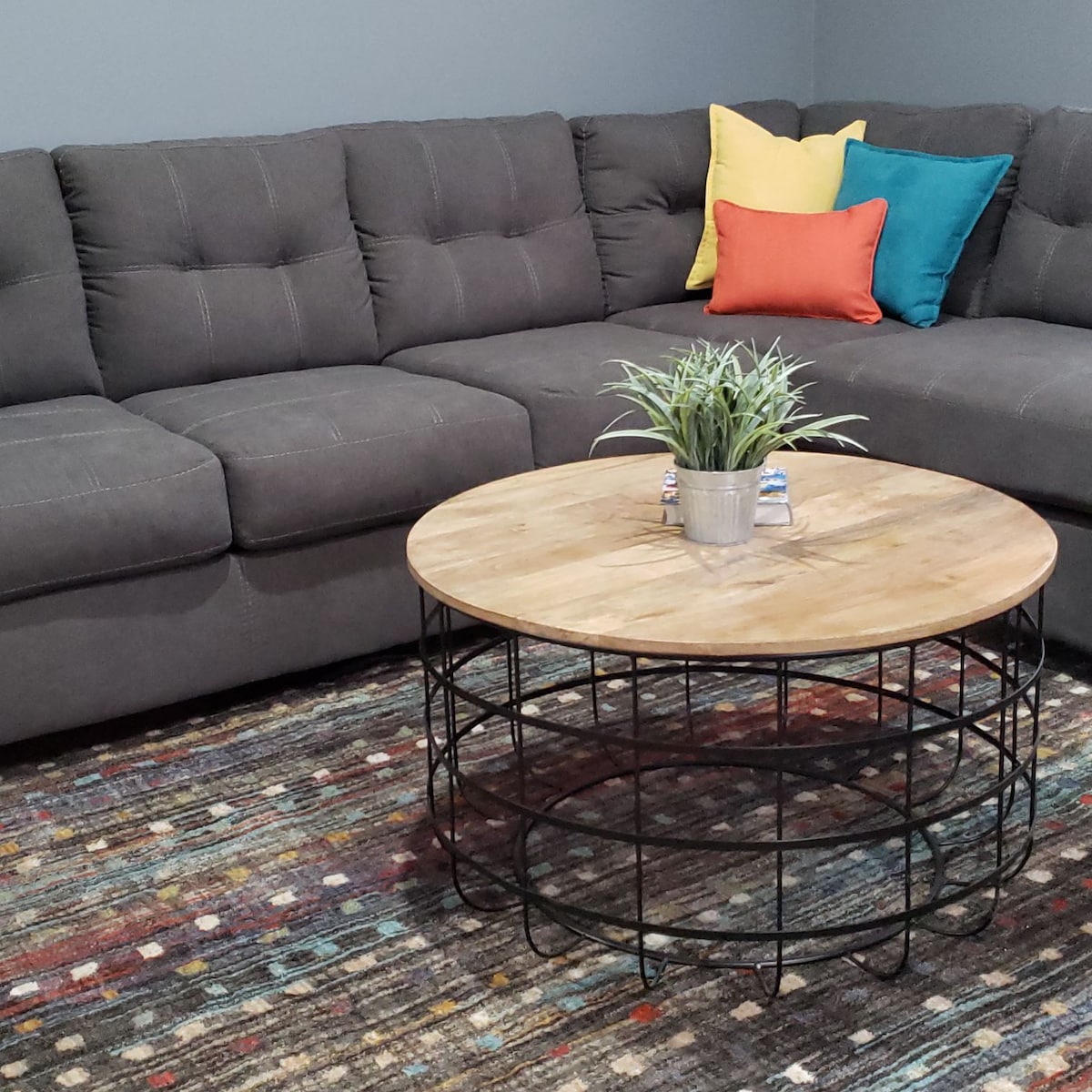 A spacious corner sofa with tufted upholstery is paired with a round wooden coffee table. Colorful cushions in yellow and teal add a vibrant touch. A potted plant sits atop the table, complemented by a textured area rug featuring a mix of colors.