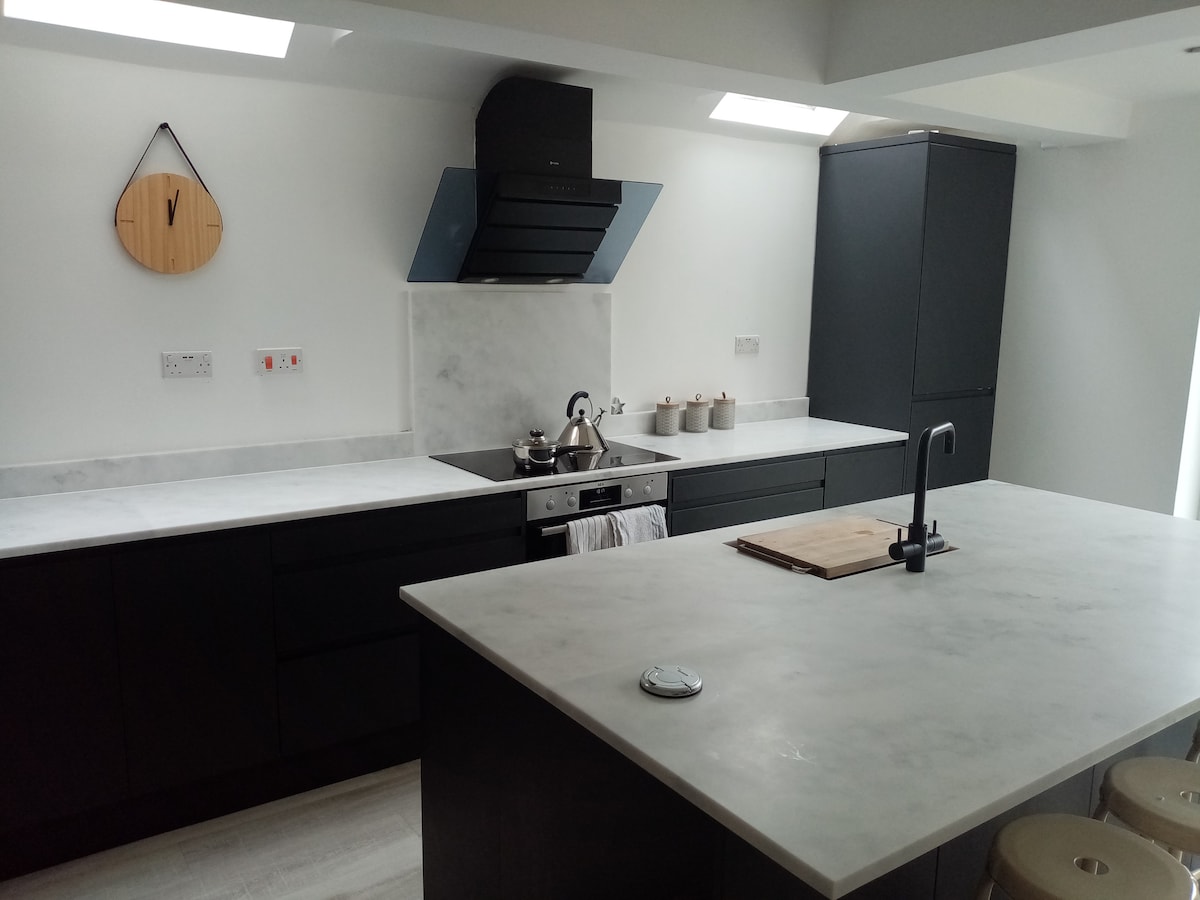 The kitchen features dark cabinetry complemented by a marble countertop. A sleek stove with a hood is visible, alongside a minimalist sink. Natural light filters in through the ceiling skylight, highlighting a clean, modern design. A chopping board and ceramic containers are placed on the countertop.